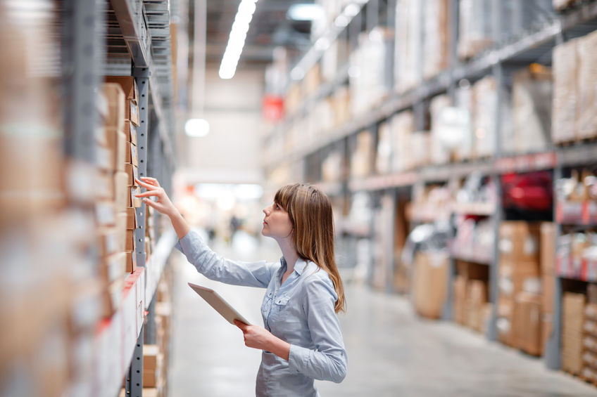 Serious staff woman wrting on notepad at supermarket Woman standing in a warehouse aisle, holding a clipboard and reaching for a box on a high shelf.